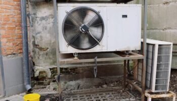 Home Exterior view of an industrial air conditioning unit with visible wear and rust, set against a shabby wall.